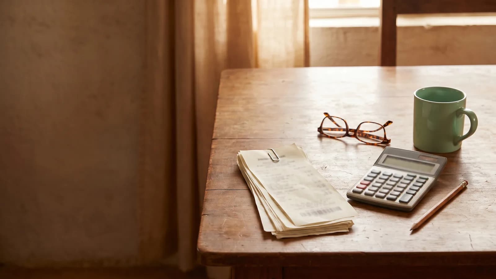 A wooden table with a clipped stack of receipts, a calculator, a pencil, a pair of glasses and a mint-green cup.